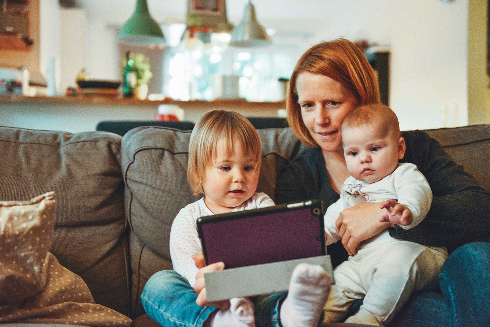 A woman looks at a tablet with a baby and toddler